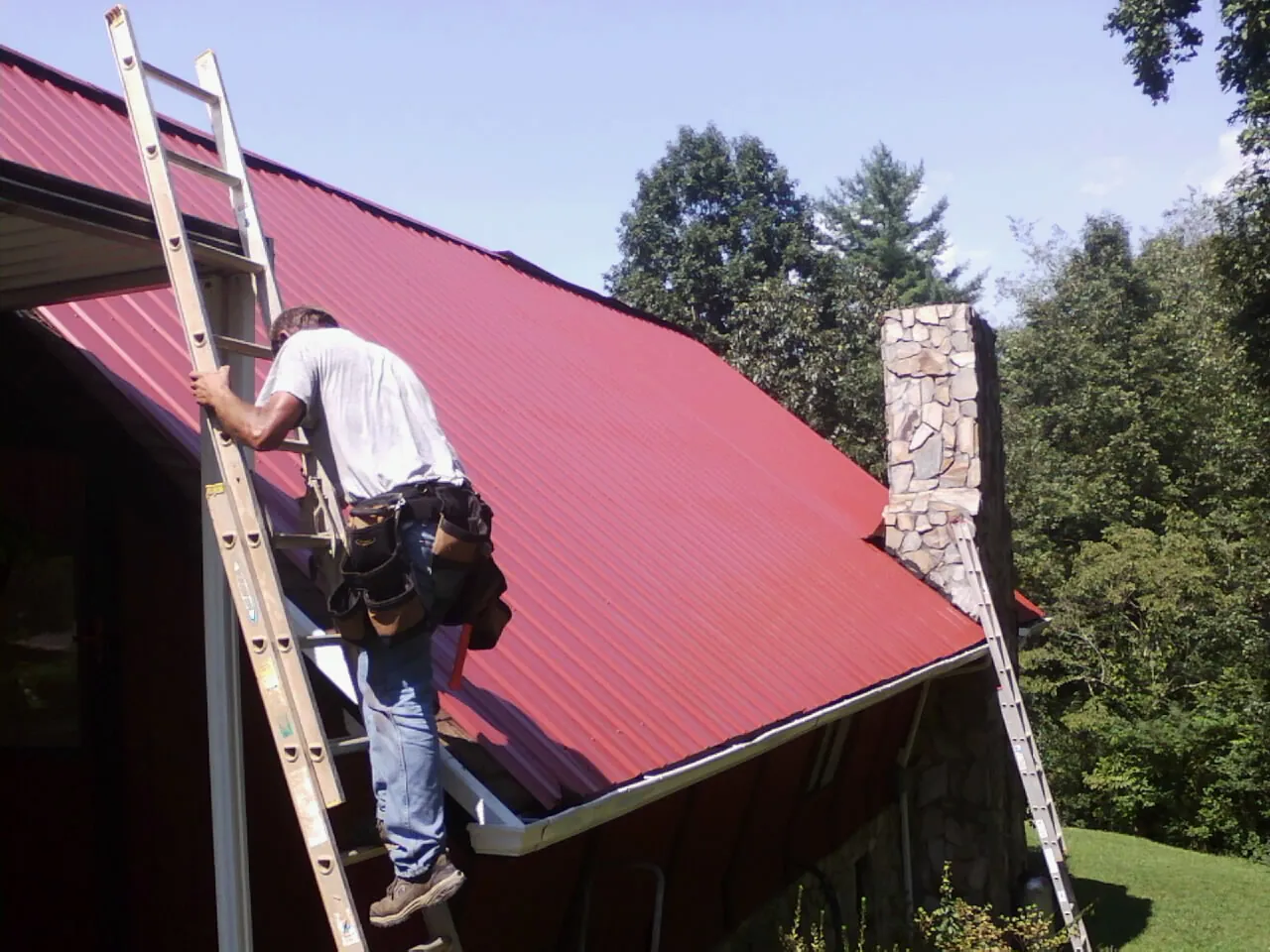 Roofer on a ladder beside a red metal roof and stone chimney — Soffit & Fascia Repair by Roofing Brothers in Pierrepont Manor